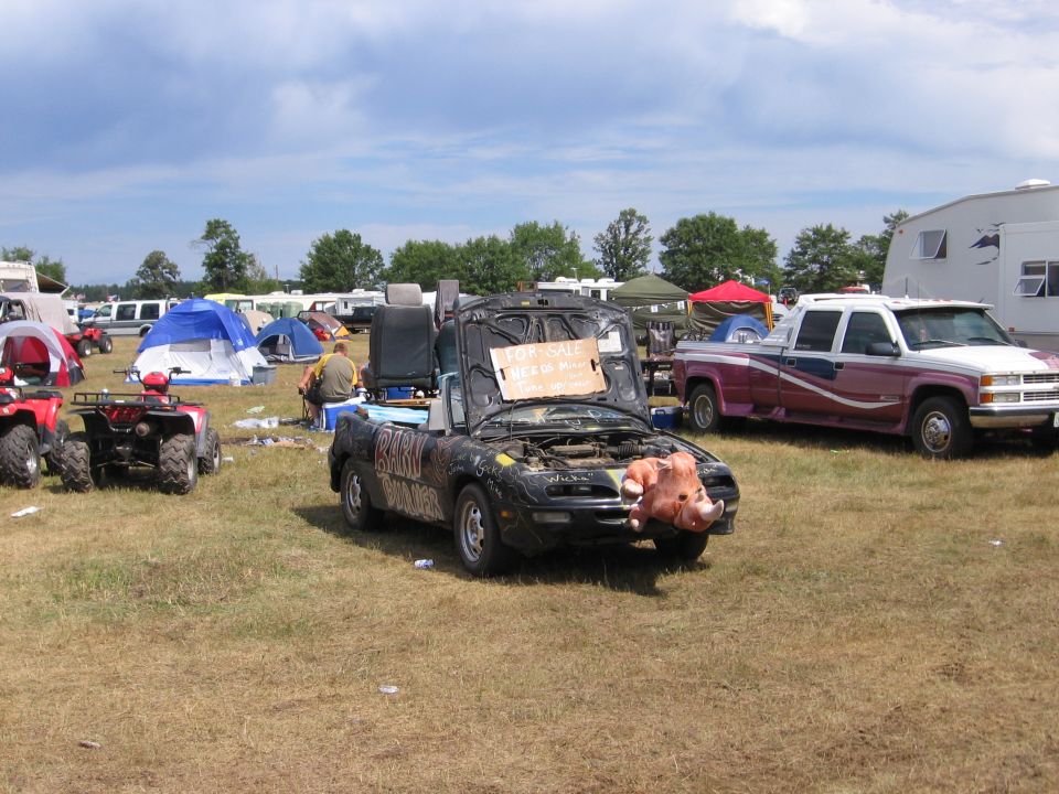 modified black convertible with pig nose, "FOR SALE" sign on hood, parked in a field with tents and ATVs