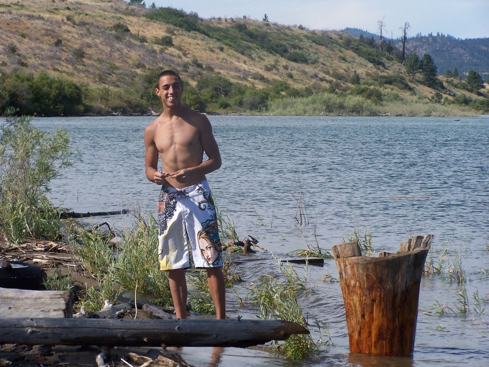 Man in patterned shorts stands in shallow water by a lake