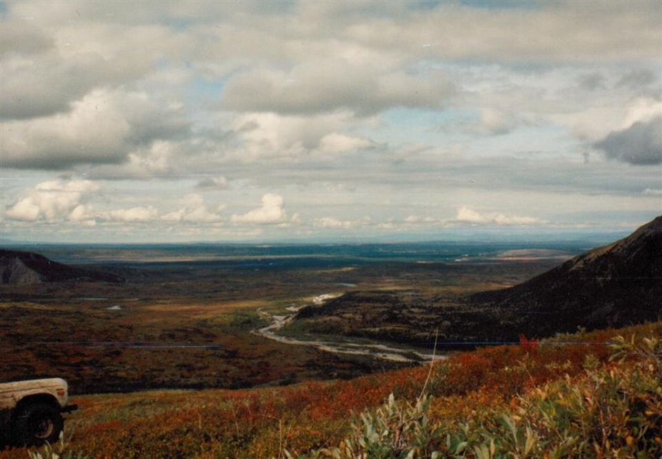 Front corner of a light-colored SUV parked on a hill overlooking a wide, open valley with a river winding through it under a cloudy sky