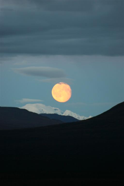 Full moon rises over snow-capped mountains and dark hills under a cloudy sky