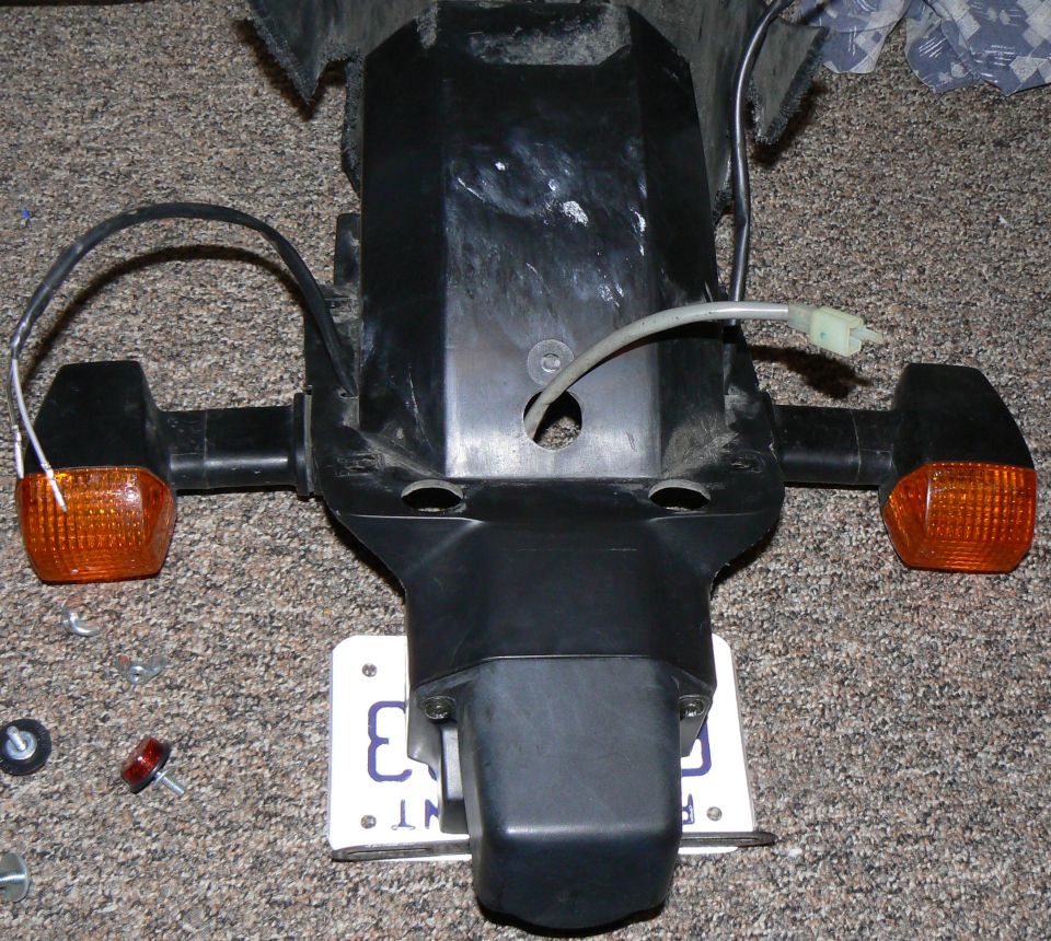 Motorcycle tail section with orange turn signals and a license plate. Some loose hardware is on the carpet
