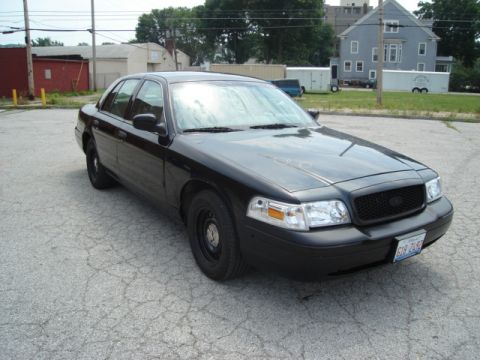 2000 Ford Crown Victoria - A black 2000 Ford Crown Victoria is parked on cracked asphalt in front of industrial buildings.