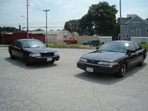 2000 Ford Crown Victoria - Two black 2000 Ford Crown Victorias are parked side-by-side in an outdoor lot under a partly cloudy sky.