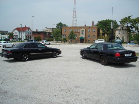 2000 Ford Crown Victoria - Two black Ford Crown Victorias are parked in a lot with buildings in the background.