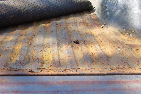 1993 Ford Bronco - The interior of a truck bed shows significant rust damage, with a dark mat rolled up in the upper left corner.