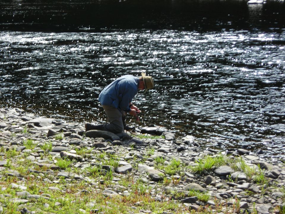 Man kneeling by a rocky riverbank, with a watermarked "SUPER MOTORS" logo in the corner