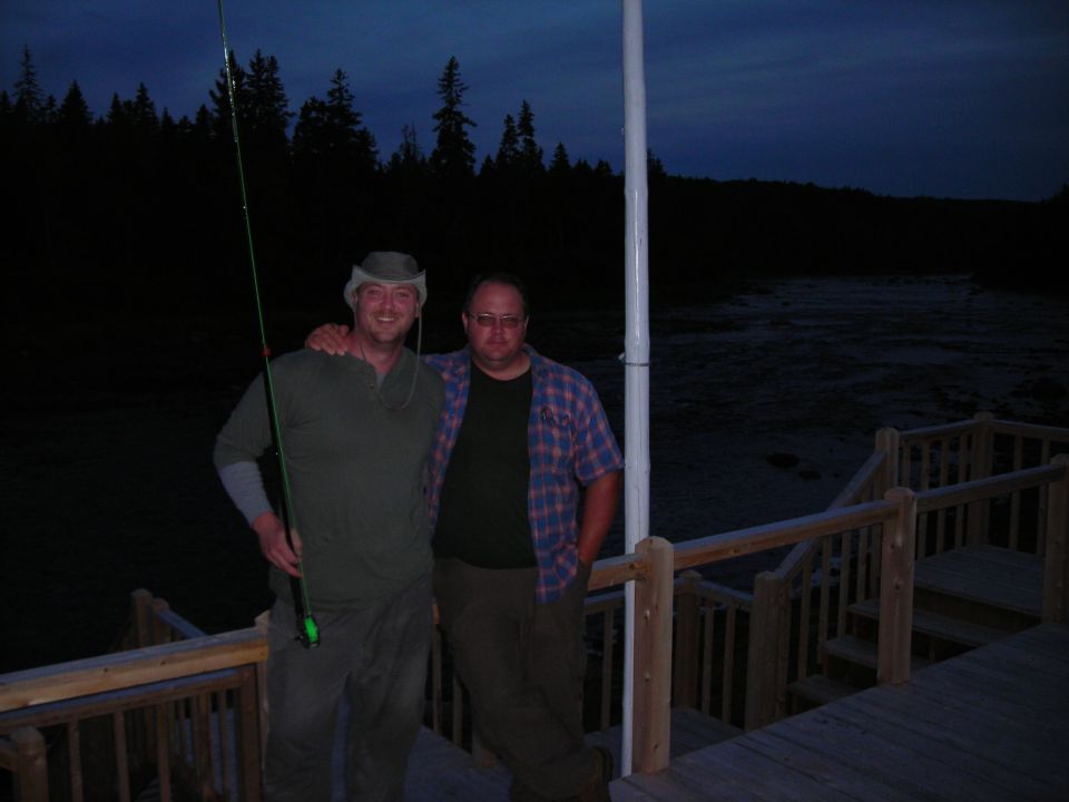 Two men stand on a wooden deck with a fishing rod, dusk scene