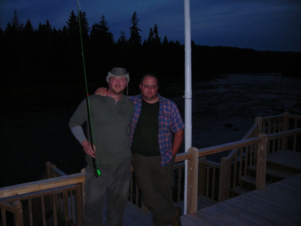 Two men stand on a wooden deck by a body of water at dusk. One man holds a fishing rod