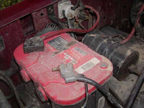 1991 Ford Bronco - A dusty Optima RedTop spiral-cell battery sits in the engine bay of a burgundy vehicle with heavy-gauge cables clamped to its posts, next t…