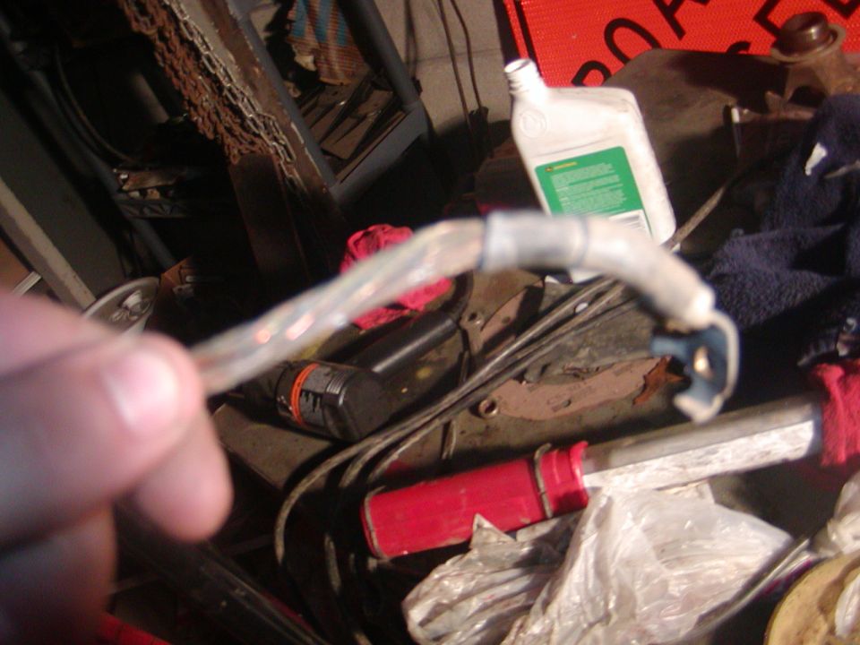 Close-up of a clear plastic hose with wires, held by a hand, with various automotive parts and a bottle of fluid in the background