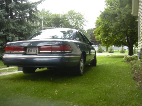 1996 Ford Crown Victoria - The rear of a dark blue sedan is visible, parked on a grassy lawn beside a house.