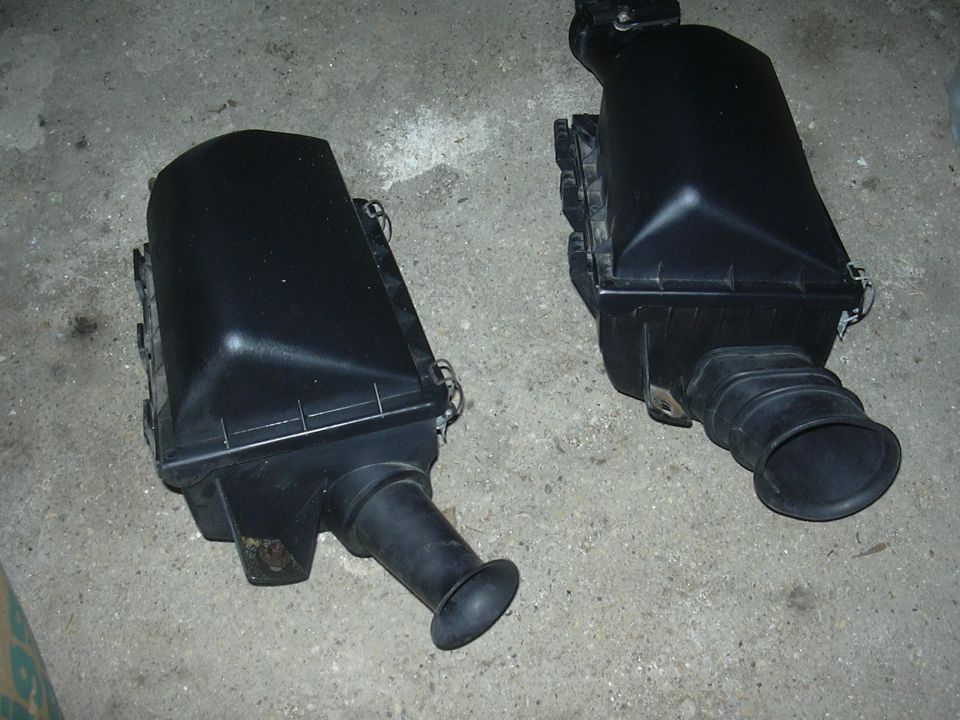 Two black plastic air filter housings on a concrete surface. One is on the left, angled slightly away. The other is on the right, closer and more head-on