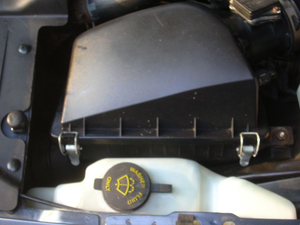 Engine bay close-up of a black airbox with spring clips above a washer fluid reservoir
