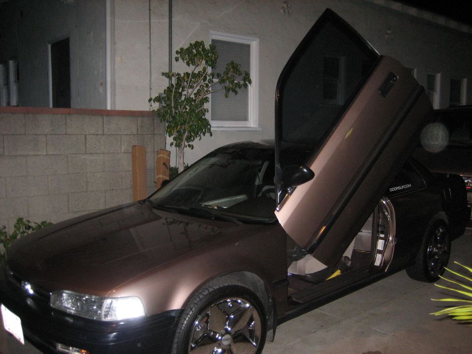 Brown sedan with doors opened upwards, parked in a driveway at night