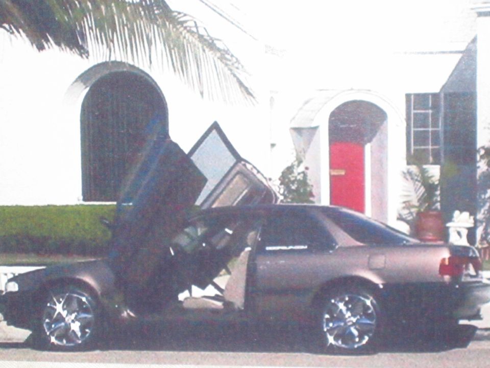 Dark coupe with doors open upwards, parked in front of a white house with a red door