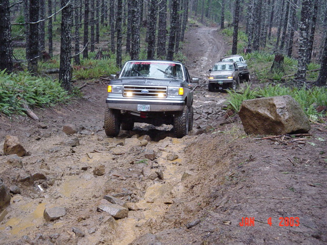 Gray Ford pickup with headlights on crawling a rocky muddy trail through dense forest