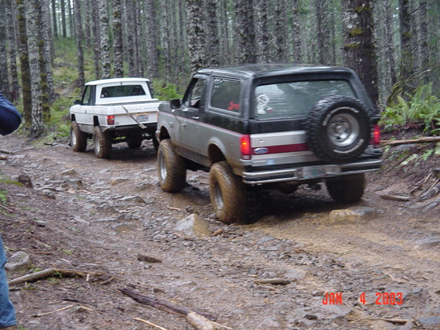 Rear view of gray Ford Bronco with spare tire towing a white pickup down a muddy forest road