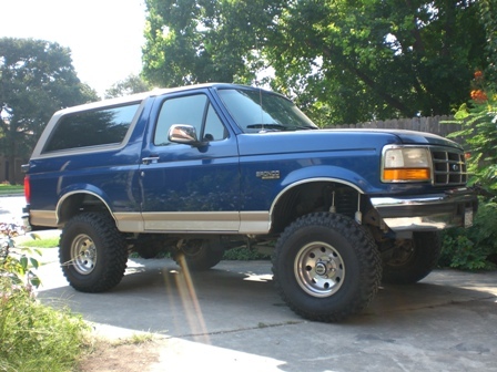 1996 Ford Bronco - A lifted blue Ford Bronco is parked on a paved surface with trees in the background.