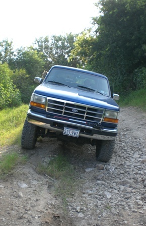 1996 Ford Bronco - A blue Ford truck is driving on a rocky, unpaved road surrounded by trees.