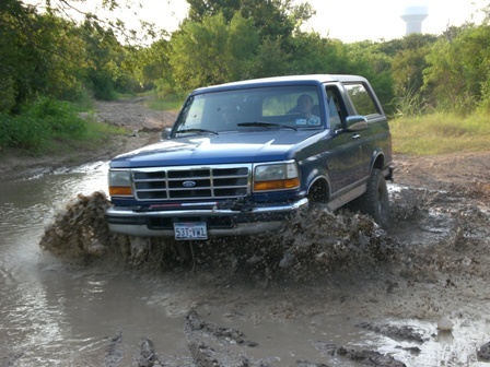 1996 Ford Bronco - A blue Ford Bronco navigates a muddy trail, kicking up water and dirt.