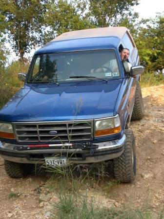 1996 Ford Bronco - A blue 1996 Ford Bronco is shown off-roading on a dirt path with a driver visible inside.