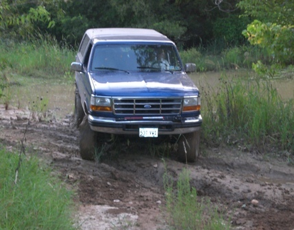 1996 Ford Bronco - A blue Ford Bronco navigates a muddy off-road path with water and vegetation surrounding it.
