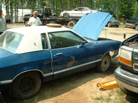 1978 Ford Mustang II - Blue coupe with white vinyl roof, hood open, parked next to a pickup truck. Another car is on a flatbed tow truck in the background.