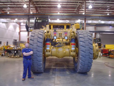 1966 International School Bus - A man stands beside a colossal yellow Caterpillar mining truck, its immense rear tires and suspension.