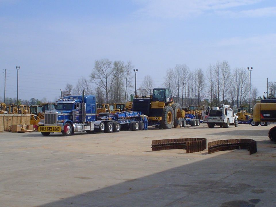 Blue semi-truck hauling a large yellow construction vehicle on a flatbed trailer. Other construction equipment visible in background