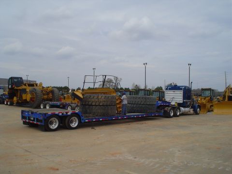 1966 International School Bus - A blue semi-truck transports heavy equipment and tires on a flatbed trailer, with other construction vehicles in the background.