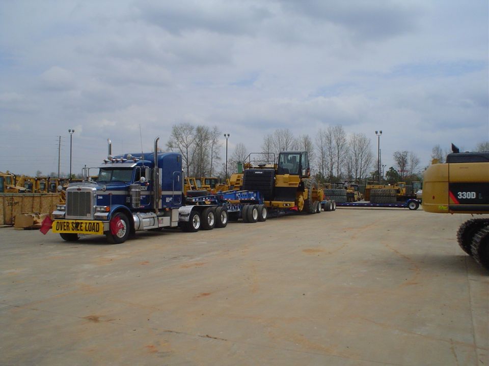 Blue Peterbilt semi-truck with "OVER SIZE LOAD" sign, hauling yellow heavy machinery on a flatbed trailer