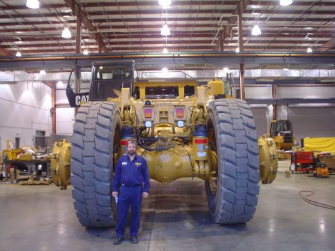 1966 International School Bus - Man in front of a large Caterpillar mining truck in a workshop. Another piece of heavy equipment is in the background.