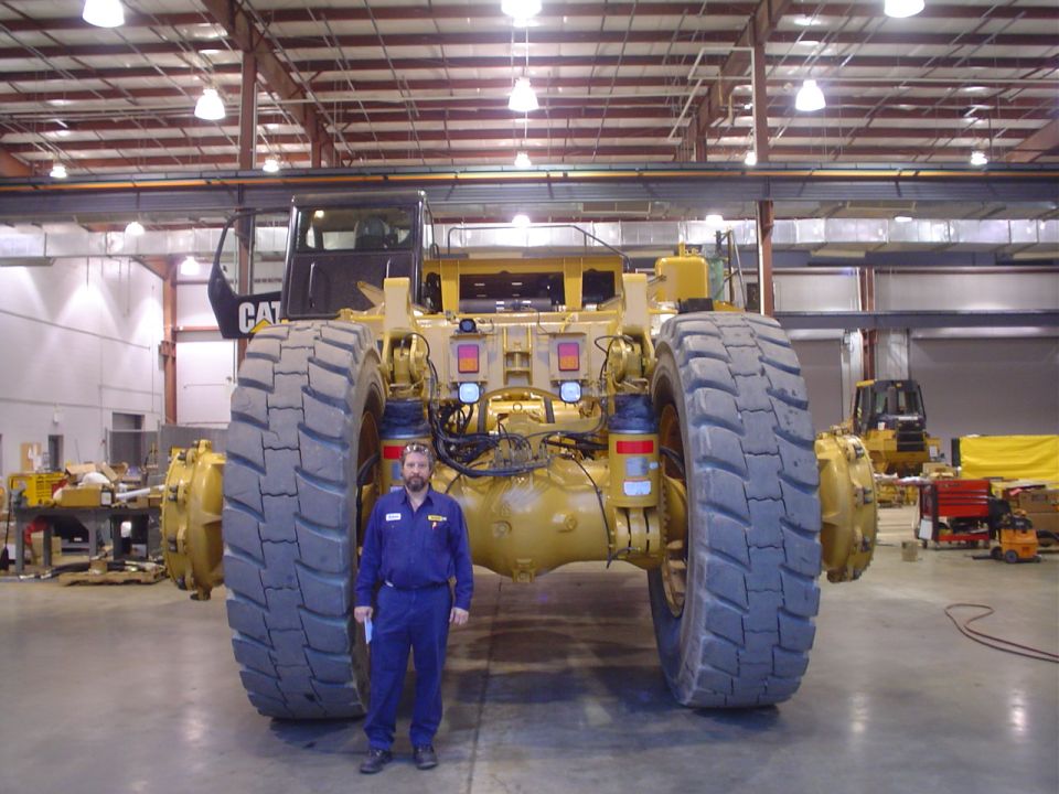 Man in blue jumpsuit stands before the massive rear tires and drivetrain of a yellow Caterpillar mining truck. Another yellow loader is visible in the background
