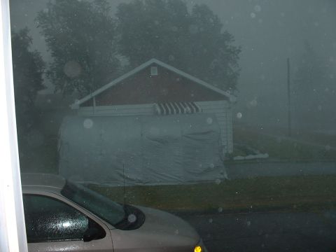 1978 Ford Mustang II - A car is seen through a window during a storm, with a house and a covered structure visible in the hazy background.