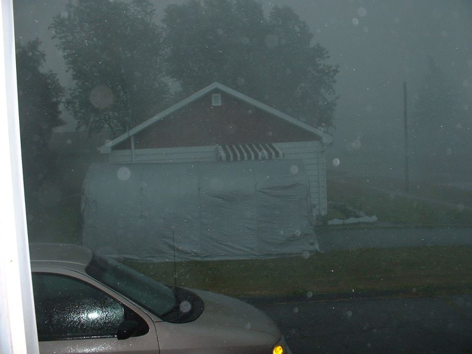 Tan sedan in driveway during heavy rainstorm, red garage with tarp behind