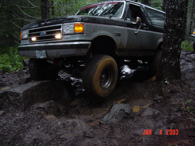 Brown Ford Bronco with headlights on climbing a wet rock obstacle, front three-quarter view