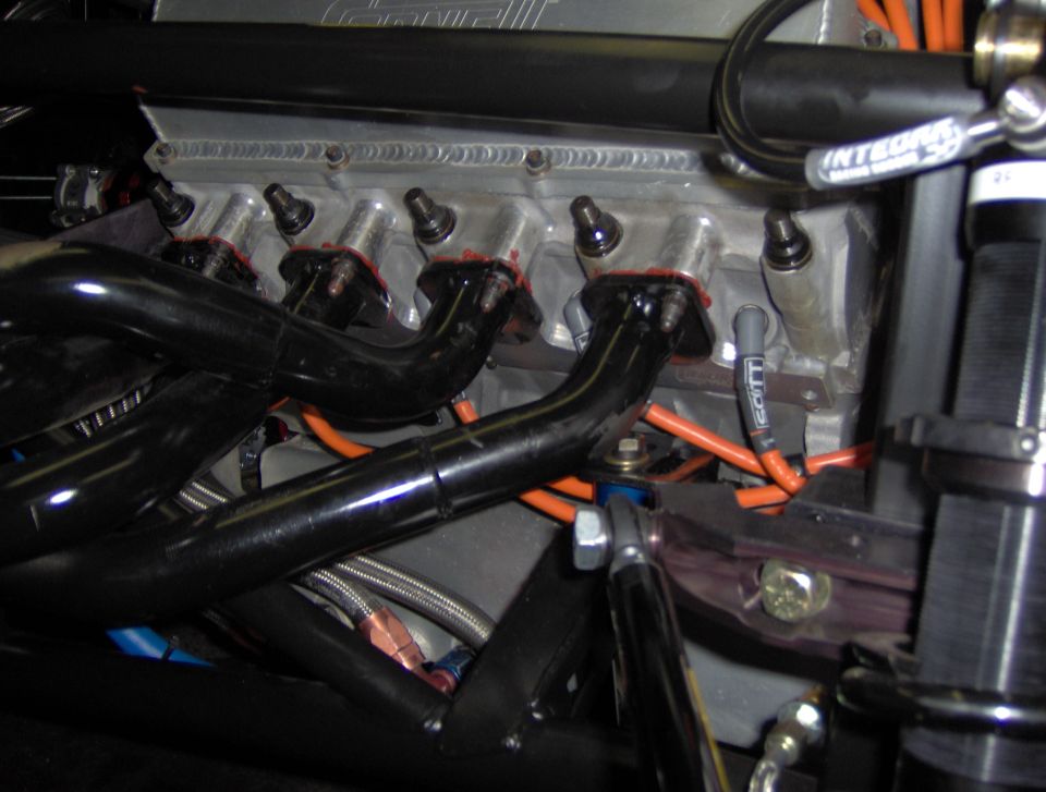 Close-up of engine with black exhaust headers, orange spark plug wires, and silver intake manifold. Some text visible on wires and a valve