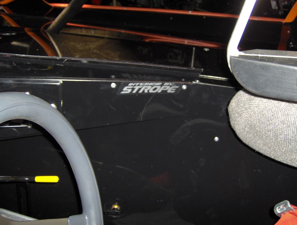 Interior of a vehicle showing a gray steering wheel, roll cage, and a badge that reads "INTERIOR BY STROPE"