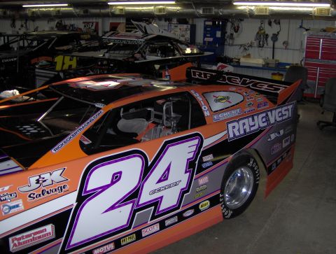 1978 Ford Bronco - A race car with sponsor decals is parked inside a garage next to another race car.