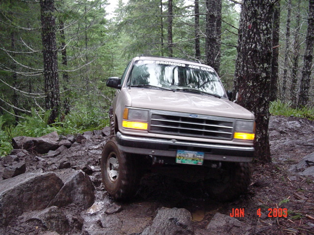 Front view of tan Ford Explorer two-door crawling over wet boulders against a tree