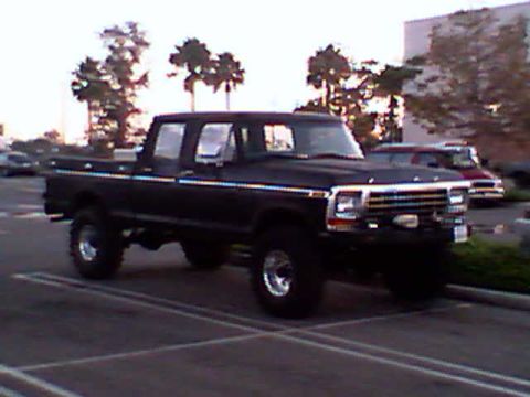 1990 Ford Bronco - A lifted black Ford F-Series pickup truck is parked in a lot with trees and a building in the background.