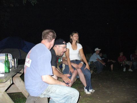 1991 Ford Probe - A group of people are sitting around a campfire at night, with tents and a picnic table in the background.