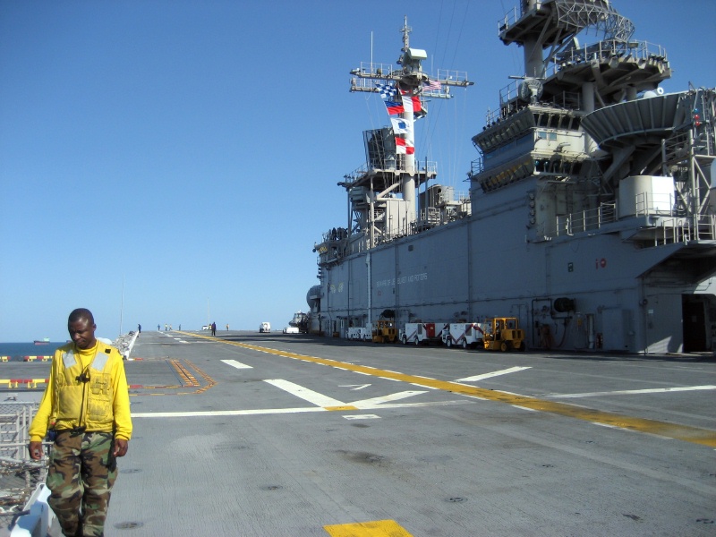 Man in yellow vest on aircraft carrier deck with ship in background