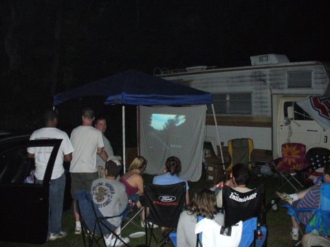 1991 Ford Probe - An outdoor gathering at night, with people watching a movie projected onto a screen near an RV.