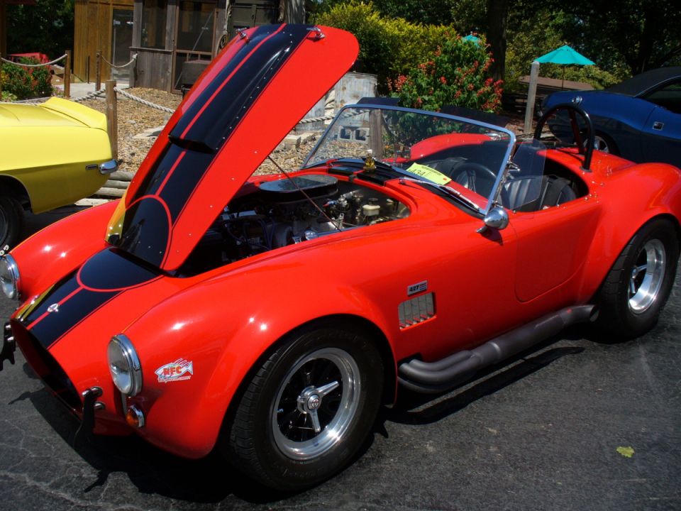 Red convertible sports car with black stripes, hood open showing engine, parked at an outdoor car show next to a yellow car