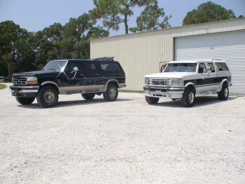 1993 Ford C-350 (Centurion Conversions) - Two Ford SUVs, a black Bronco and a white F-250 Centurion conversion, are parked outside a metal building.