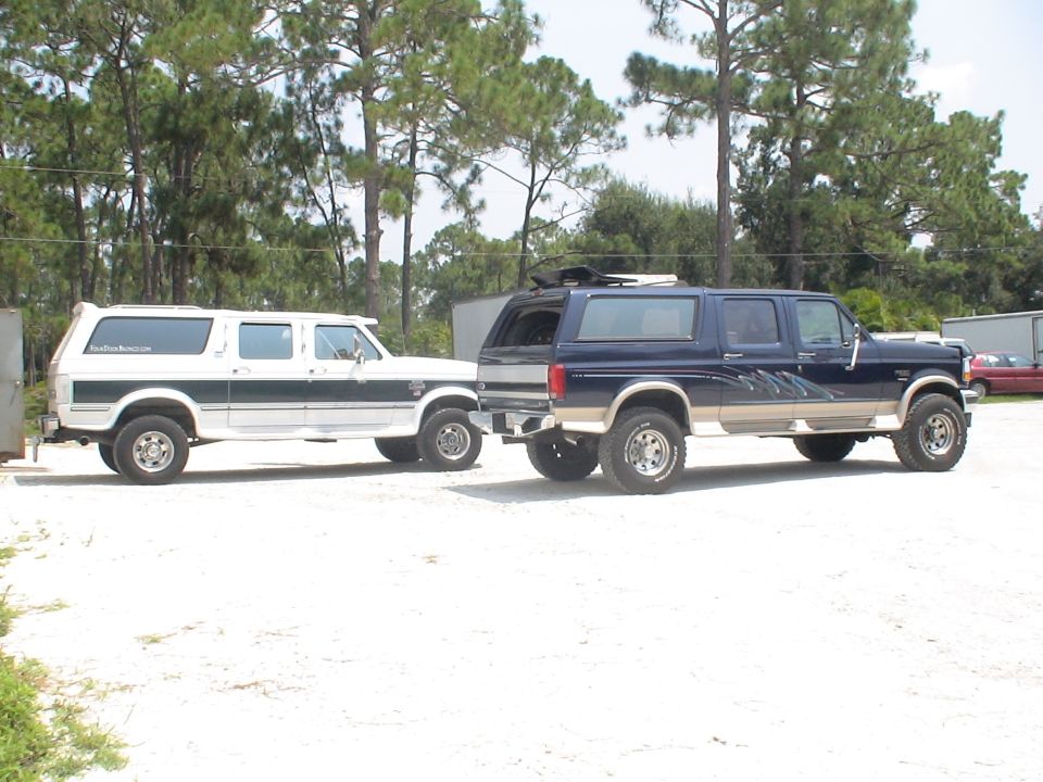 Two Ford C-350 Centurion Conversion SUVs parked side-by-side. The white one is on the left, the blue one on the right. Both are lifted
