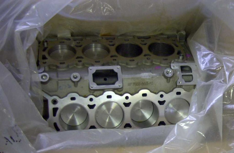 Engine block with six cylinders, partially covered in plastic wrap. Text "MY 7000-005-P2" is visible