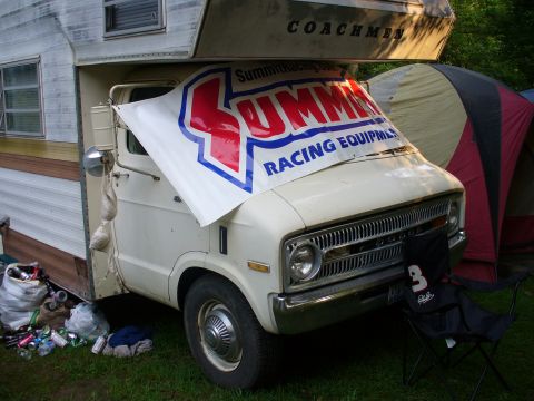 1991 Ford Probe - An old RV is parked on grass, partially covered by a large racing equipment banner.