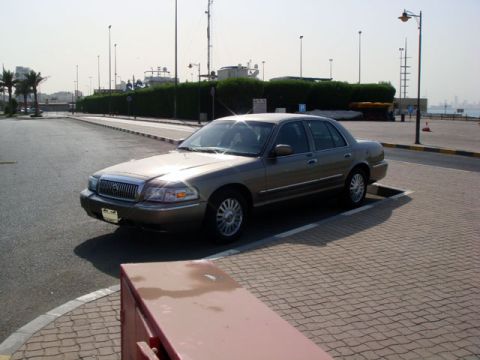 2007 Mercury Grand Marquis - A tan/gold four-door sedan parked at an angle in an empty paved lot, viewed from the front three-quarter left with sun glare on the windshi…
